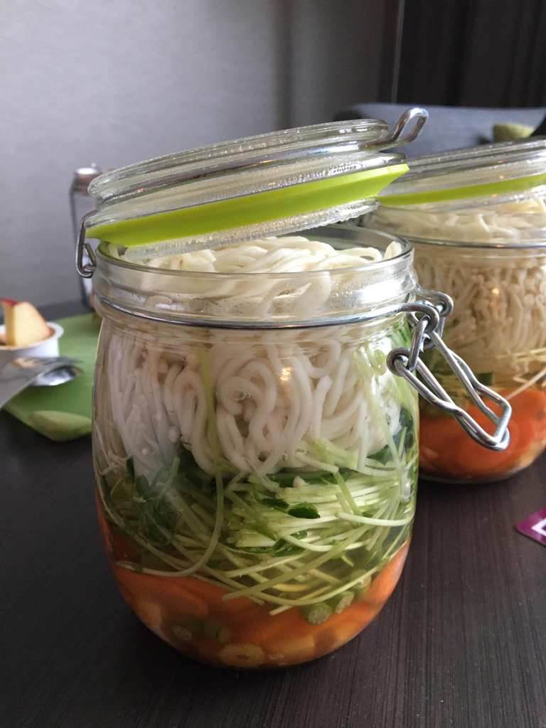 A glass jar layered with vegetables, greens, and noodles