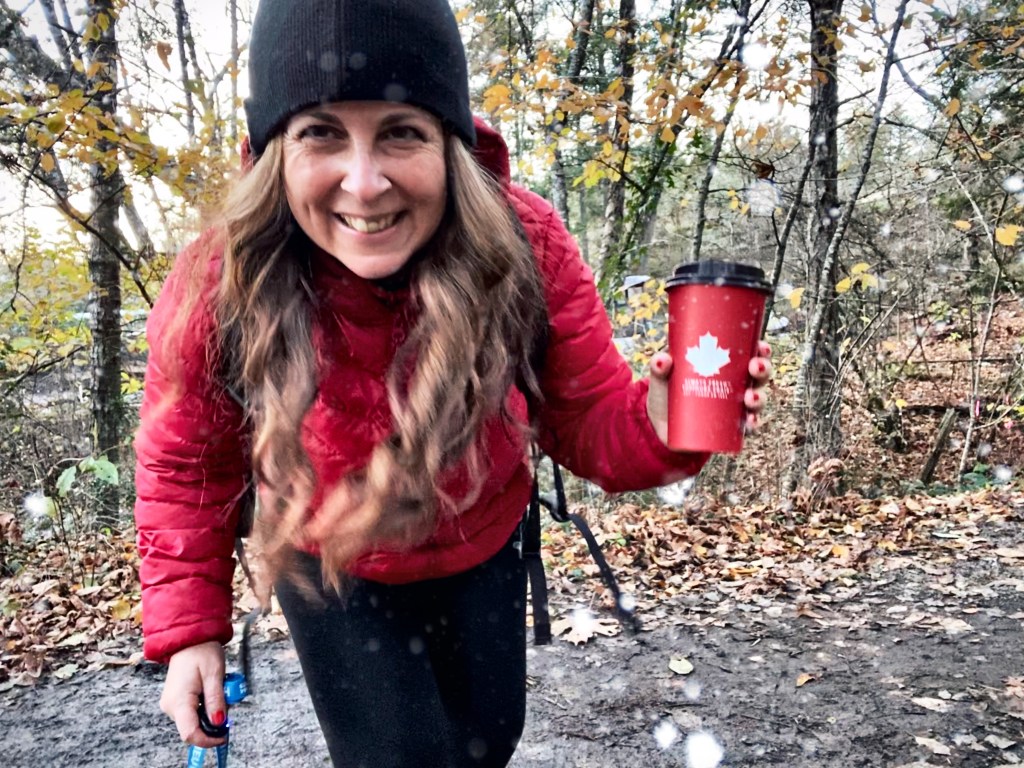 Charlotte Millington smiling on a wooded trail, wearing a red jacket and holding a travel coffee cup